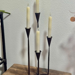 Puede incluir: Un candelabro de hierro forjado con cuatro velas cónicas de color crema. El candelabro tiene un acabado marrón oscuro y un diseño curvo único. Las velas tienen un efecto de parpadeo, agregando un ambiente cálido.