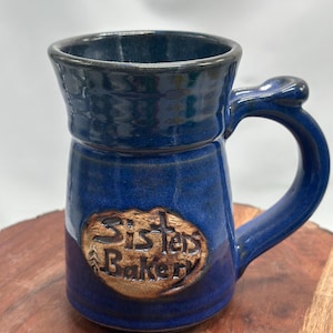 Könnte beinhalten: Eine blaue Keramiktasse mit dunkelblauem Rand und Henkel. Die Tasse trägt den Text "Sister's Bakery" in einem erhabenen, braunen ovalen Design auf der Vorderseite. Die Tasse hat eine glänzende Oberfläche und einen abgerundeten Henkel.