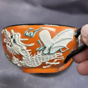 Op de afbeelding: Oranje theekopje met een zwart handvat en een wit drakenontwerp. De draak is in reliëf en heeft blauwe accenten.
