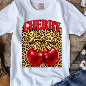 Op de afbeelding: Wit T-shirt met een grafische afbeelding met het woord "CHERRY" in het rood, boven een luipaardprint strik en twee glanzende rode kersen. Het shirt wordt getoond met laarzen en denim.
