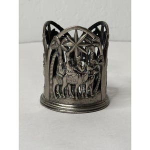 Può includere: Un portabicchieri in metallo color argento a tema religioso. Il design presenta tre figure a cavallo di cammelli, palme e una stella. Il portabicchieri ha un design traforato e una base circolare.