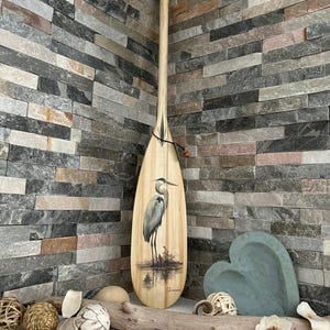 May include: A wooden paddle featuring a painting of a heron. The paddle is light brown with a dark brown accent and a black cord. The heron is grey and white, standing in water. The paddle is displayed against a stone wall.