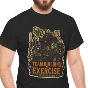 May include: A black t-shirt featuring a graphic illustration of a necromancer and skeletons with the text "TEAM BUILDING EXERCISE" and "ANIMATE DEAD" in orange and green lettering. The design includes a sword, a spellbook, and skulls.