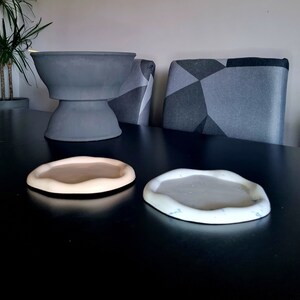 Peut inclure: Deux sous-verres ronds en céramique, l'un de couleur beige clair et l'autre de couleur gris clair. Les sous-verres ont un bord ondulé et organique. Les sous-verres sont posés sur une table noire.