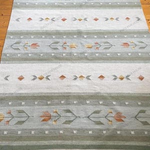 Puede incluir: Alfombra rectangular con un patrón geométrico en tonos verdes, blancos y naranjas. La alfombra presenta un diseño repetitivo de formas florales y de diamantes estilizados, con un borde con flecos. La alfombra se muestra sobre un suelo de madera.