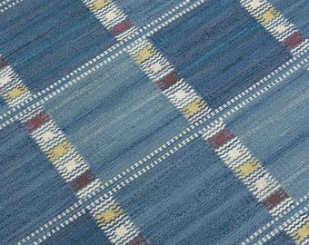 Op de afbeelding: Een geweven textiel met een blauw ruitpatroon. Het ontwerp bestaat uit grote blauwe vierkanten gescheiden door kruisende witte, gele en donkerrode gestreepte banden. De stof heeft een zichtbaar geweven textuur met diverse blauwtinten.