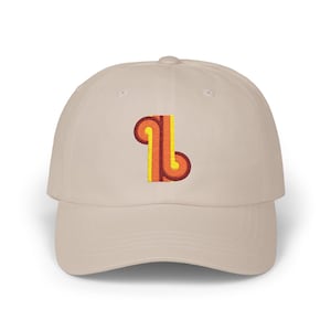 May include: A beige baseball cap with a retro-style embroidered design. The design features a vertical, stylized letter "L" in shades of orange and yellow. The cap has a curved brim and a classic six-panel construction.