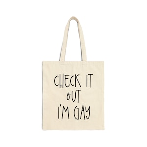 Può includere: Una borsa tote in tela beige con testo nero che dice "CHECK IT OUT I'M GAY".