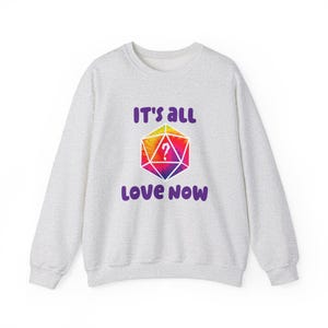 Può includere: Una felpa grigio chiaro con la scritta "IT'S ALL LOVE NOW" in viola. Al centro è presente una forma geometrica colorata con un punto interrogativo. La forma presenta un gradiente arcobaleno di colori tra cui giallo, arancione, rosa e viola.