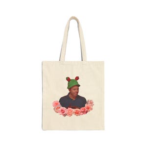 Può includere: Borsa tote color crema con un uomo che indossa un cappello verde con bacche rosse e circondato da rose rosa. L'uomo indossa una camicia blu scuro. La borsa ha due manici.