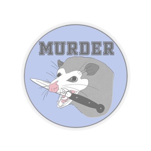 以下が含まれることがあります： ナイフを口にくわえた漫画のオポッサムが描かれた丸いステッカー。オポッサムはグレーと白で、ピンクの鼻と口をしています。「MURDER」の文字が、薄い青色の背景の上に、オポッサムの上に灰色のブロック体で印刷されています。