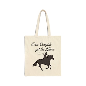Può includere: Una borsa tote beige con una silhouette nera di una cowgirl a cavallo e il testo "Even Cowgirls get the Blues".