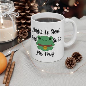 以下が含まれることがあります： 緑色のカエルのイラストと「Magic Is Real And So Is My Frog」という文字が書かれた白いセラミック製のマグカップです。