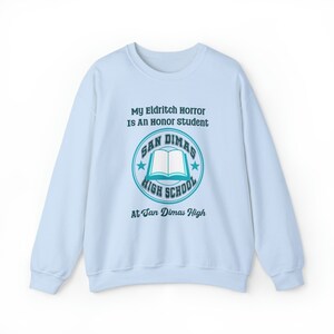 May include: Light blue sweatshirt with a graphic print of a book and the text "My Eldritch Horror Is An Honor Student" and "San Dimas High School" with a star on each side of the text. The text "At San Dimas High" is printed below the graphic.