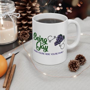 Può includere: Tazza in ceramica bianca con la scritta "Being Gay" in verde, accompagnata da un'illustrazione di uva viola. La frase "When you're here you're family" è stampata sotto. La tazza è su una superficie grigio chiaro.