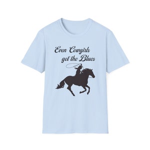 Può includere: Maglietta blu chiaro con una silhouette nera di una cowgirl a cavallo. Il testo "Even Cowgirls get the Blues" è stampato sulla maglietta.
