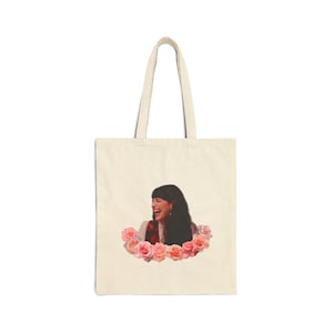 Può includere: Borsa tote color crema con una persona sorridente circondata da rose rosa e pesca. La borsa ha due manici ed è ideale per trasportare oggetti. Il design è semplice e la borsa è realizzata in tessuto dai toni neutri.