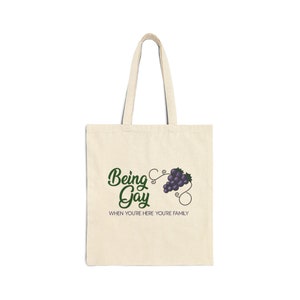 Peut inclure: Un sac fourre-tout beige avec le texte "Being Gay When You're Here You're Family" et une vigne verte avec des raisins violets.