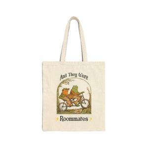 Puede incluir: Una bolsa de tela beige con un gráfico de dos ranas montando una bicicleta. El texto "And They Were Roommates" está impreso en la bolsa.