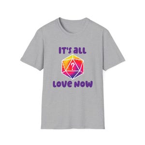 Può includere: Una maglietta grigia con la scritta "IT'S ALL LOVE NOW" in viola. La maglietta presenta una forma geometrica colorata con un punto interrogativo al centro, utilizzando sfumature di giallo, arancione, rosa e viola.
