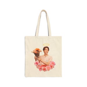 Può includere: Borsa tote color crema con una grafica di una persona che tiene in mano un bouquet di fiori arancioni e rosa, circondato da rose rosa. La persona ha un'aureola sopra la testa. La borsa ha due manici.