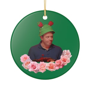 Può includere: Ornamento rotondo in ceramica verde. Presenta un uomo con un cappello verde con dettagli di fragole e una polo blu, circondato da rose rosa. Un filo d'oro è attaccato per appenderlo.
