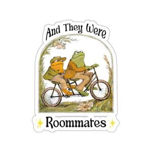 Puede incluir: Una pegatina con dos ranas de dibujos animados montadas en un tándem. Una rana es verde, la otra naranja. El texto "And They Were Roommates" está arqueado sobre las ranas, con un borde decorativo y estrellas.