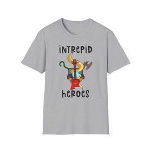 Può includere: Una t-shirt grigia melange con una grafica di una chitarra, un serpente, uno staff, un'ascia e un globo. Il testo "intrepid heroes" è stampato sotto la grafica.