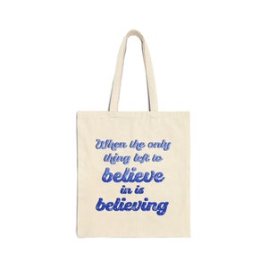 Può includere: Una borsa tote beige con testo blu che recita "When the only thing left to believe in is believing".