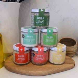 Puede incluir: Colección de seis tarros de cristal de sales marinas, incluyendo Hierbas Asadas, Océano Puro, Coco Lima, Ahumada, Pimienta y Chile y Ajo. Cada tarro tiene una tapa blanca, una etiqueta y un acento verde o rojo. Los tarros están colocados sobre una tabla de madera.