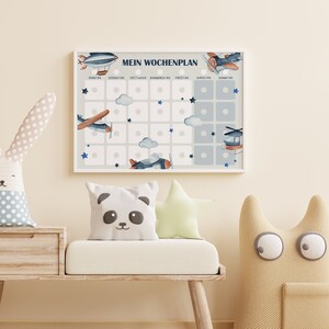 Op de afbeelding: Een wit ingelijst schema met een blauw en wit vliegtuigthema. Het schema is gelabeld "Mein Wochenplan" en heeft zeven kolommen die zijn gelabeld met de dagen van de week in het Duits. Elke kolom heeft vijf vierkanten met een witte cirkel in het midden. Er zijn wolken en sterren op het schema.
