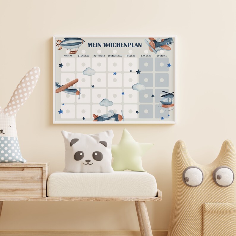 Op de afbeelding: Een wit ingelijst schema met een blauw en wit vliegtuigthema. Het schema is gelabeld "Mein Wochenplan" en heeft zeven kolommen die zijn gelabeld met de dagen van de week in het Duits. Elke kolom heeft vijf vierkanten met een witte cirkel in het midden. Er zijn wolken en sterren op het schema.