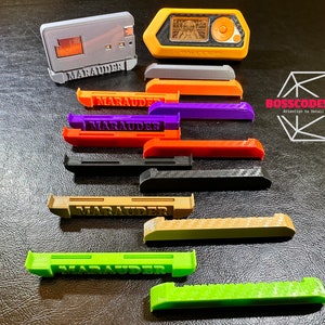Op de afbeelding: Een verzameling 3D-geprinte kunststof onderdelen in verschillende kleuren, waaronder grijs, oranje, paars, rood, zwart en groen. De onderdelen zijn allemaal gelabeld met het woord "MARAUDER".