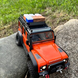 Peut inclure: Un Land Rover Defender jouet orange avec un porte-bagages de toit noir et une barre lumineuse. Le porte-bagages est charg&eacute; de plusieurs petites bo&icirc;tes et d'une caisse en bois.