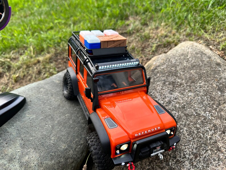 Peut inclure: Un Land Rover Defender jouet orange avec un porte-bagages de toit noir et une barre lumineuse. Le porte-bagages est charg&eacute; de plusieurs petites bo&icirc;tes et d'une caisse en bois.