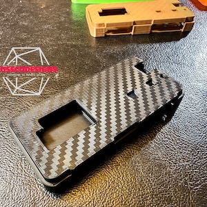 Pode incluir: Uma capa de telefone preta com um padrão de fibra de carbono. A capa tem um recorte para a lente da câmera e um recorte para a porta de carregamento.