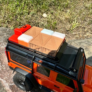 Peut inclure: Un camion jouet noir et orange avec un porte-bagages noir sur le toit. Le porte-bagages est charg&eacute; d'une caisse en bois brune, de deux bo&icirc;tes en plastique blanches et d'un r&eacute;cipient en plastique rouge.