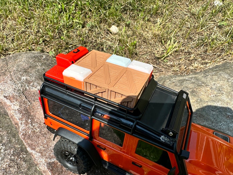 Peut inclure: Un camion jouet noir et orange avec un porte-bagages noir sur le toit. Le porte-bagages est charg&eacute; d'une caisse en bois brune, de deux bo&icirc;tes en plastique blanches et d'un r&eacute;cipient en plastique rouge.