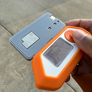 Pode incluir: Um dispositivo eletrônico cinza e laranja com uma tela que exibe uma imagem de desenho animado. O dispositivo tem um botão no lado e a palavra "RAZER" impressa no lado. O dispositivo está aberto para revelar um painel traseiro cinza com um código QR e texto.