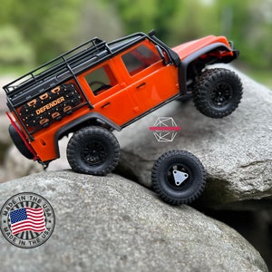 May include: Orange toy Land Rover Defender with a black roof rack and spare tire. The toy car is on a rock with a "Made in the USA" sticker.