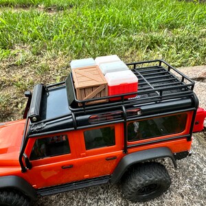 Peut inclure: Camion jouet orange avec un porte-bagages de toit noir. Le porte-bagages est charg&eacute; d'une caisse en bois et de trois contenants en plastique. Deux des contenants sont blancs et un est rouge.