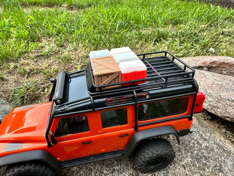 Peut inclure: Camion jouet orange avec un porte-bagages de toit noir. Le porte-bagages est charg&eacute; d'une caisse en bois et de trois contenants en plastique. Deux des contenants sont blancs et un est rouge.