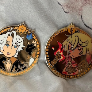 May include: Two round, gold-colored enamel pins. One features a cartoon character with white hair, blue eyes, and a black outfit. The other pin shows a cartoon character with blonde hair and a red and yellow flame. Both pins have a gold border.