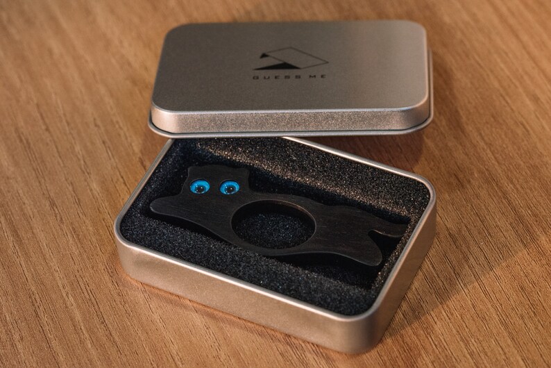 Pu&ograve; includere: Un anello di legno nero a forma di gatto con occhi blu, in una scatola argentata con la scritta "BUERR ME" sul coperchio.