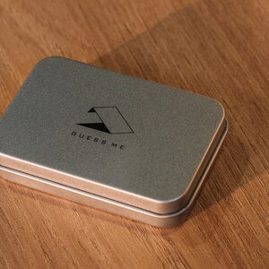 Pu&ograve; includere: Una scatola di metallo argentata con un logo nero che dice "GUESS ME" e una forma geometrica.
