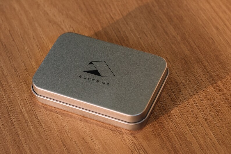 Pu&ograve; includere: Una scatola di metallo argentata con un logo nero che dice "GUESS ME" e una forma geometrica.