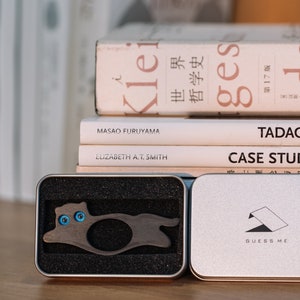 Pu&ograve; includere: Un porta anelli in legno a forma di gatto con occhi blu, in una scatola nera e argentata. La scatola ha la scritta "Guess Me" stampata.