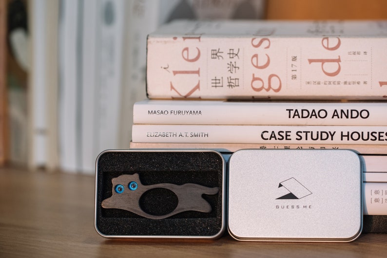 Pu&ograve; includere: Un porta anelli in legno a forma di gatto con occhi blu, in una scatola nera e argentata. La scatola ha la scritta "Guess Me" stampata.