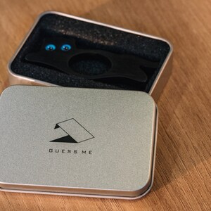 Pu&ograve; includere: Una scatola di metallo argentata con le parole "GUESS ME" stampate sul coperchio. La scatola &egrave; aperta e contiene un oggetto di metallo nero con due occhi blu.