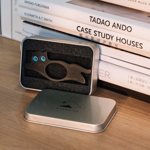 Pu&ograve; includere: Un portachiavi in metallo nero a forma di gatto con occhi blu, in una scatola argentata con interno in schiuma nera. La scatola &egrave; aperta e il portachiavi &egrave; visibile. La scatola &egrave; appoggiata su una superficie di legno.